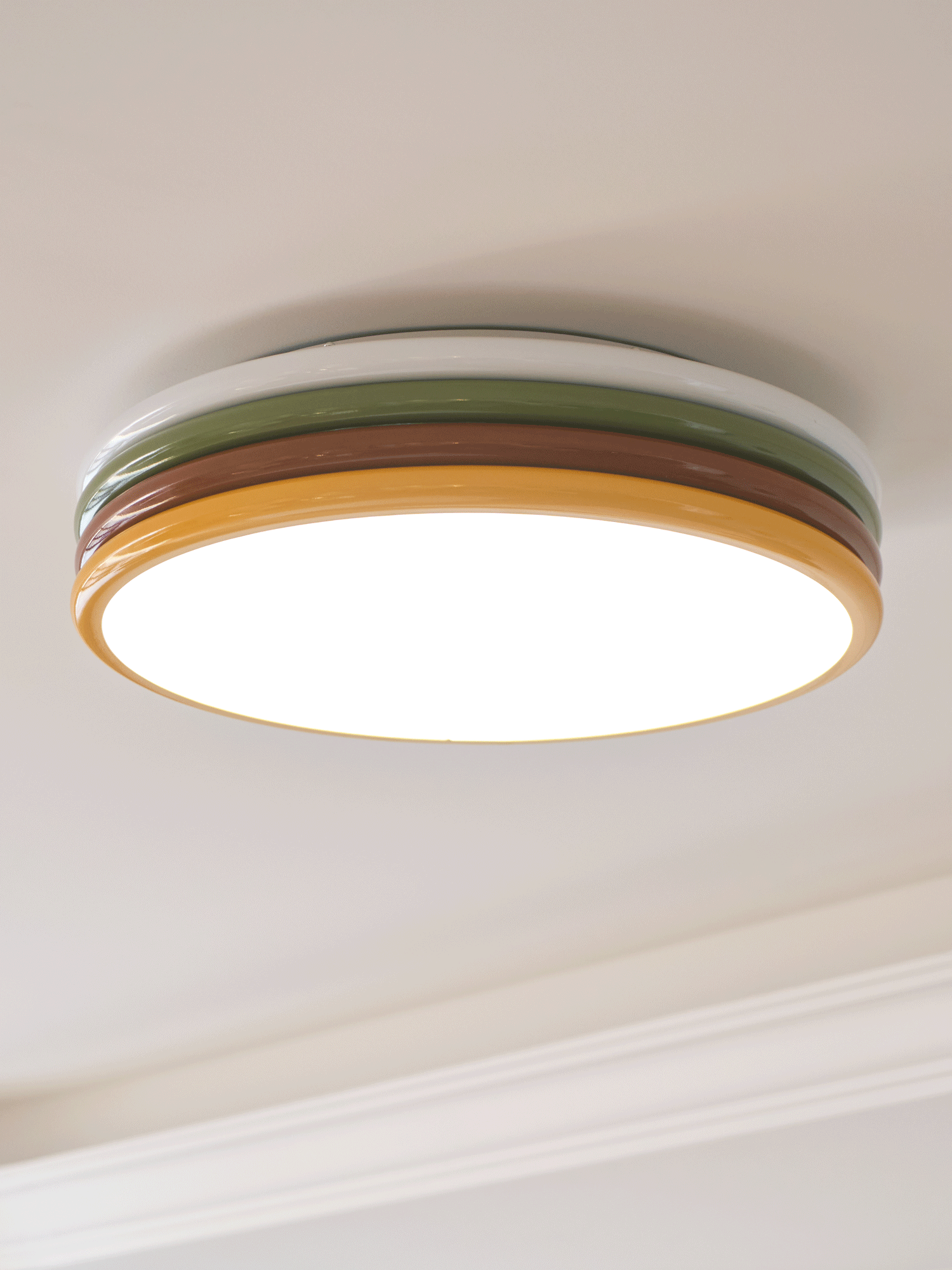 Lit Clérachic ceiling light with a bright diffuser and thin yellow rim, seen from below beside a white cornice.