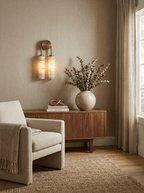 Clérachic wall sconce glowing above a walnut sideboard with a dried-flower vase, a boucle armchair and a knitted throw beside it.