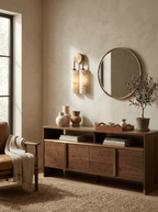 Clérachic sconce lit above a walnut sideboard and beside a round brass-framed mirror, with an olive tree in a small pot.