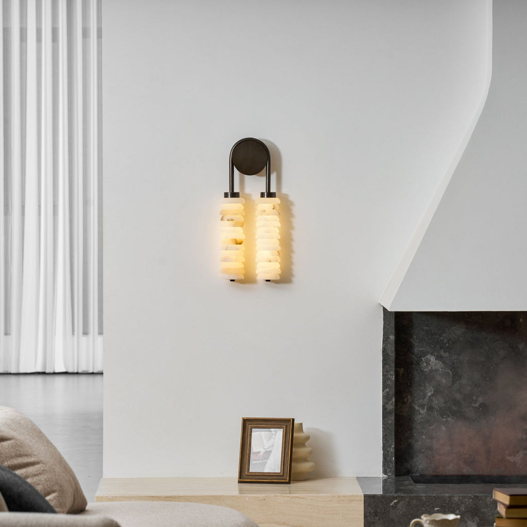 Clérachic alabaster sconce lit on a white wall beside a plastered fireplace, with a small framed picture on the ledge below.