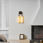Clérachic alabaster sconce lit on a white wall beside a plastered fireplace, with a small framed picture on the ledge below.