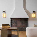 Two lit Clérachic sconces on either side of a white fireplace hood, above a round table with stacked books and a boucle pouffe.