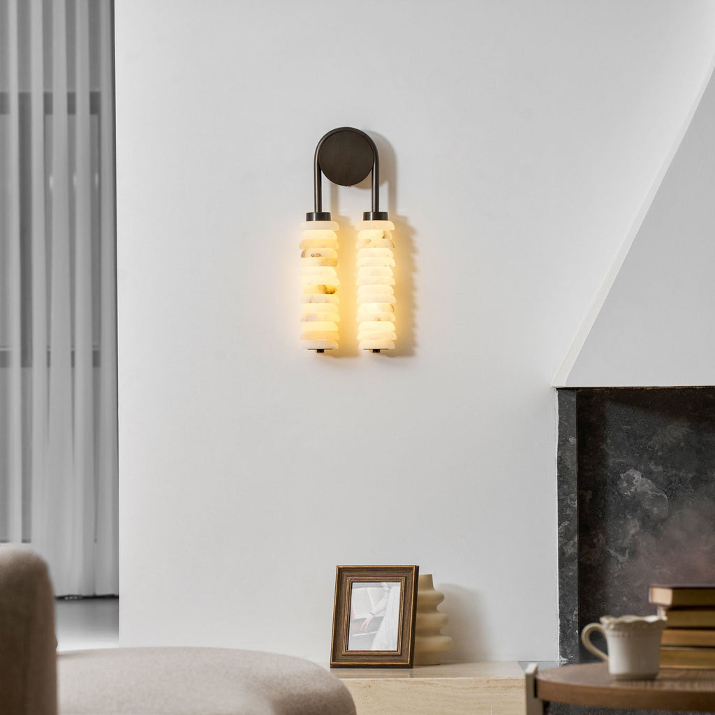 Clérachic sconce glowing amber-white on a plain wall, with a sheer curtain on the left and a framed photo on the hearth below.