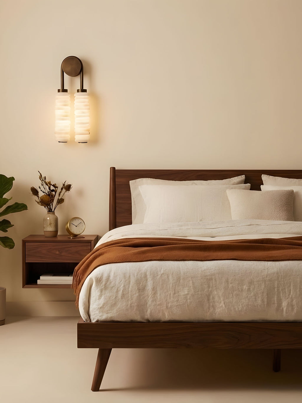 Clérachic sconce lit above a dark wood bed with rust-coloured throw and white linen, against a warm sand-coloured wall.