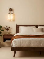 Clérachic sconce lit above a dark wood bed with rust-coloured throw and white linen, against a warm sand-coloured wall.