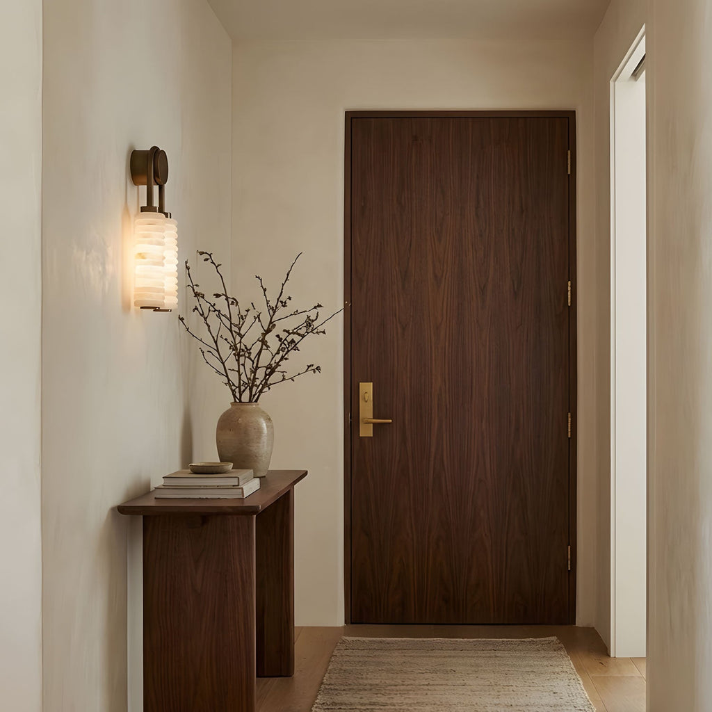 Clérachic sconce lit in a hallway beside a walnut door, above a slim console with a vase of dried branches.
