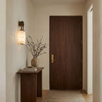 Clérachic sconce lit in a hallway beside a walnut door, above a slim console with a vase of dried branches.
