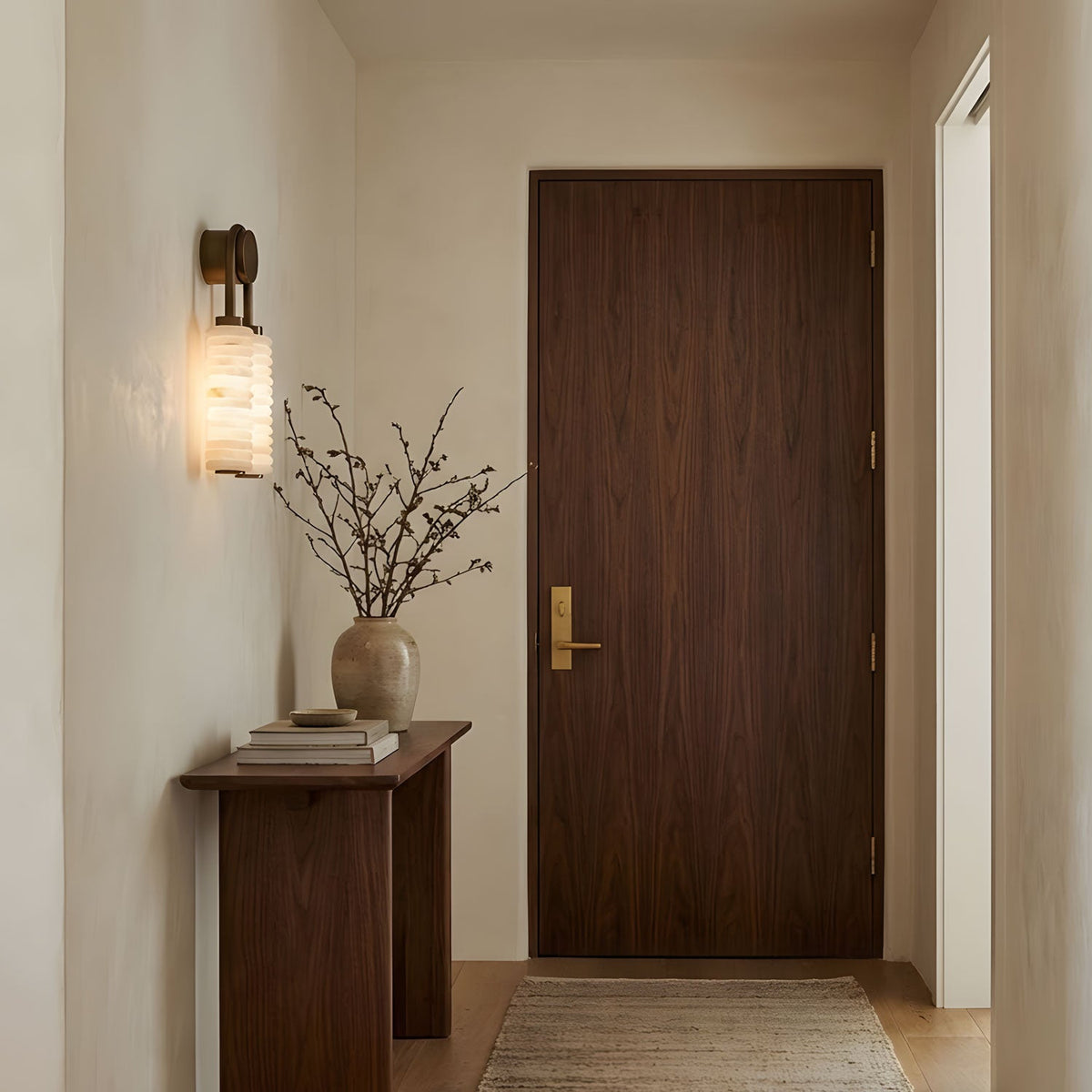 Clérachic sconce lit in a hallway beside a walnut door, above a slim console with a vase of dried branches.