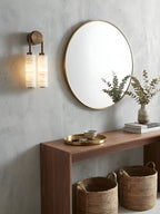 Clérachic sconce lit beside a round brass-framed mirror, above a walnut console with two woven baskets and a eucalyptus vase.
