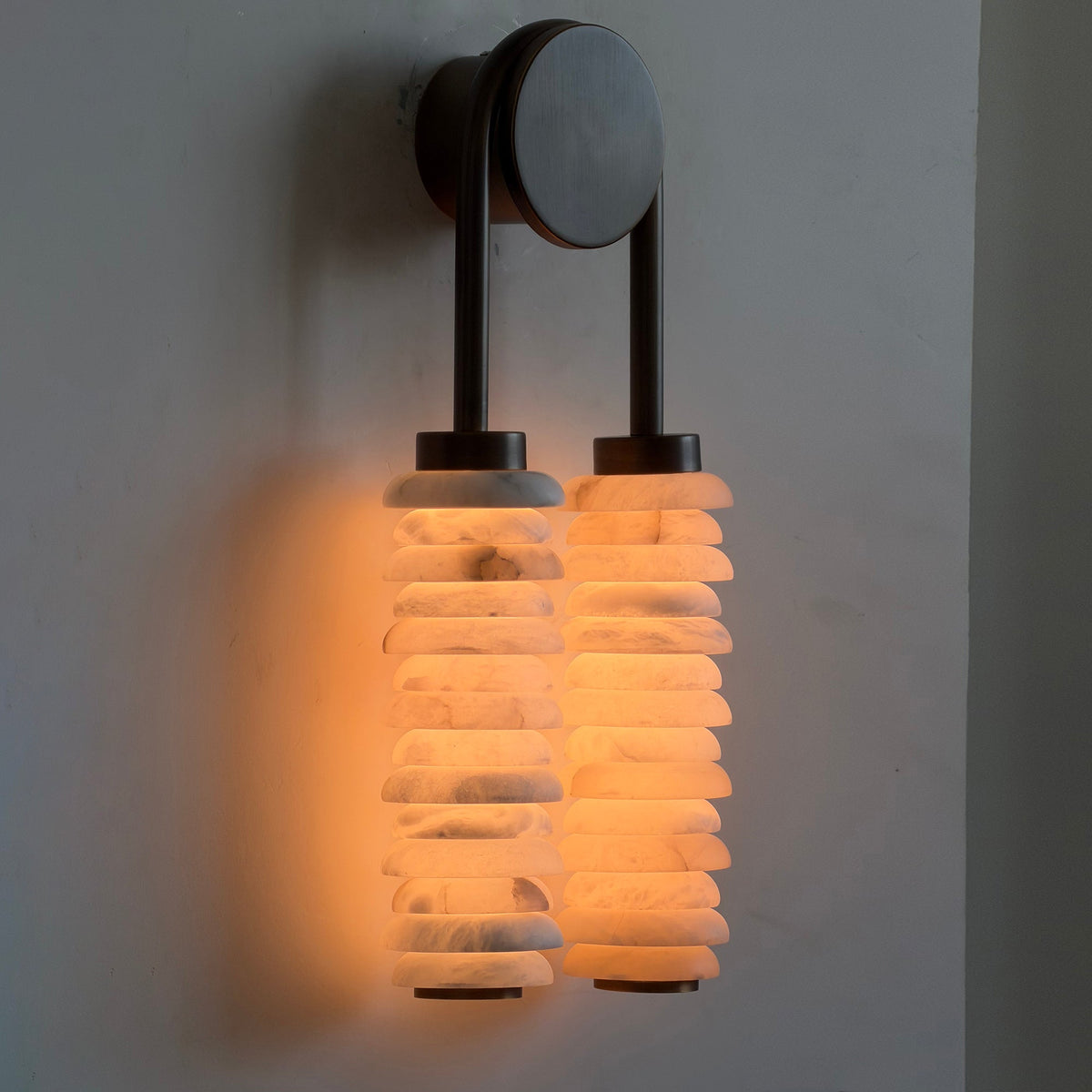 Lit Clérachic sconce on a grey wall, its alabaster discs glowing orange-amber and the veining showing through the stone.