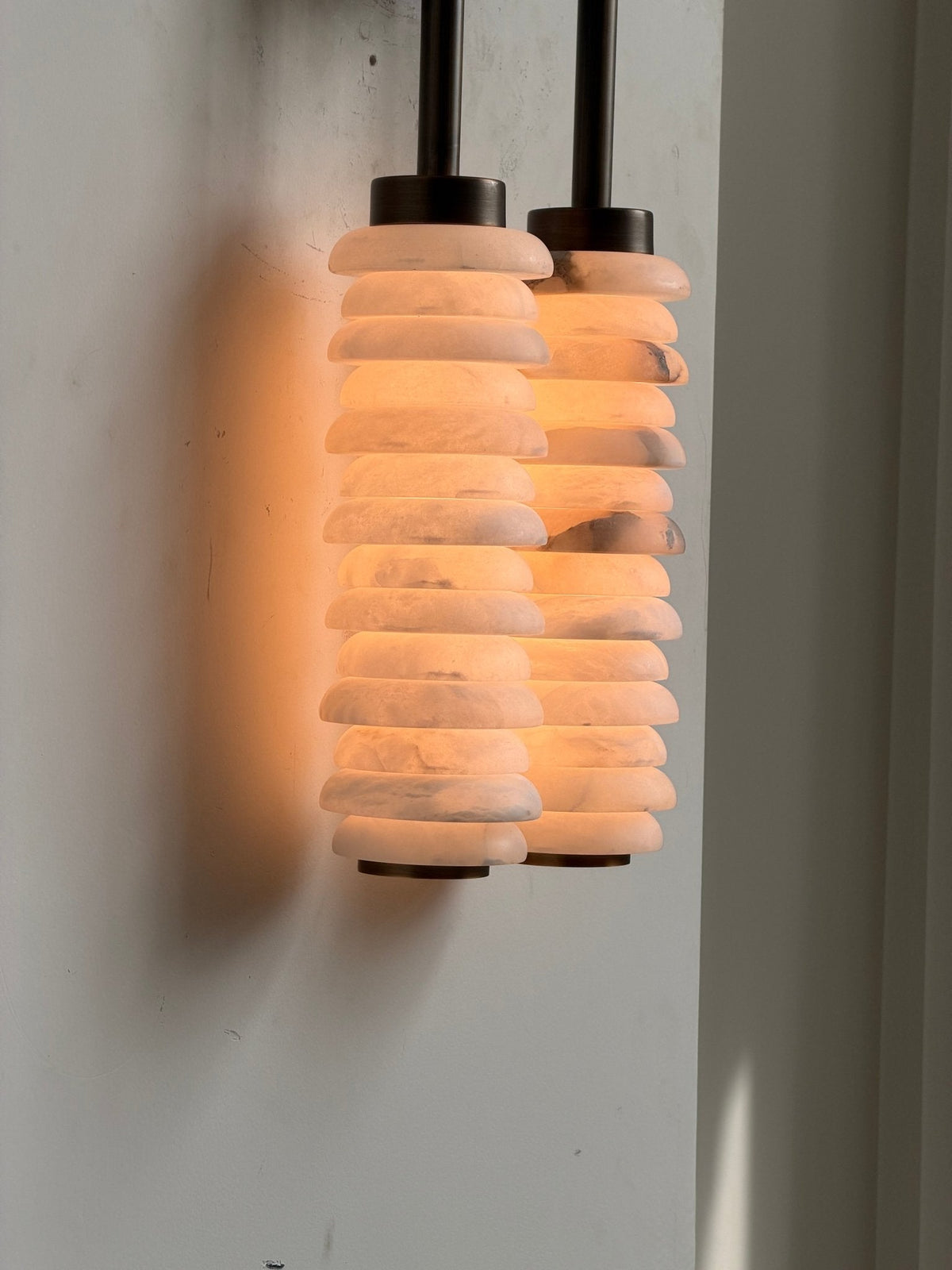 Lit Clérachic sconce on a plain wall, orange light glowing through the stacked alabaster discs and casting a soft shadow.