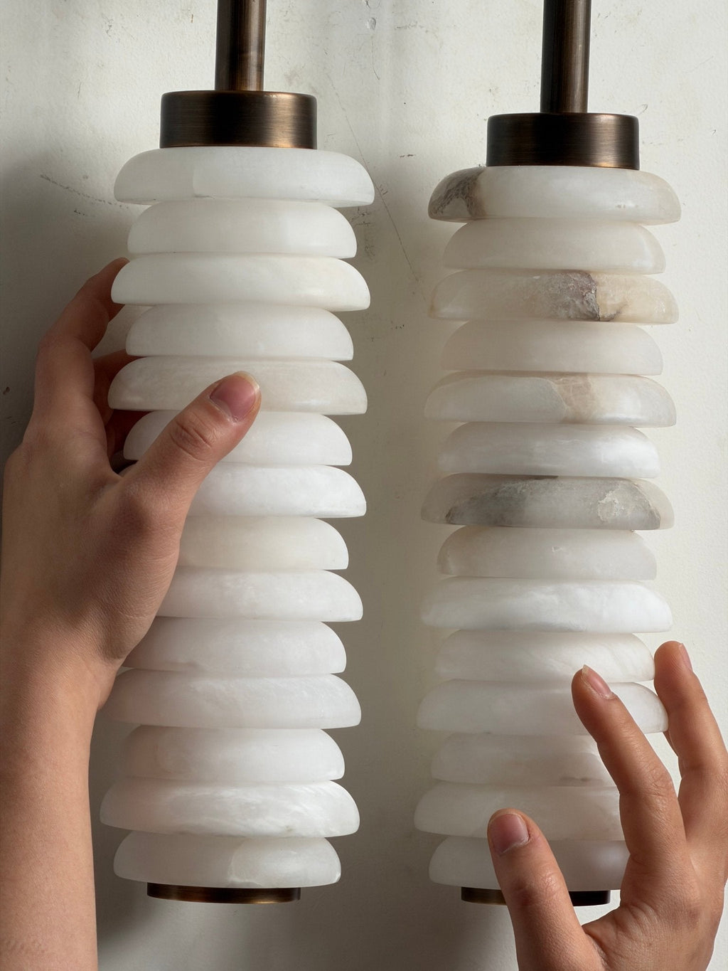 Hands steadying the two unlit alabaster columns of a Clérachic sconce, the milky white discs stacked beneath bronze caps.