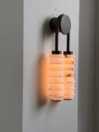 Lit Clérachic sconce on a white wall panel, its two alabaster columns glowing amber beneath a dark round backplate.