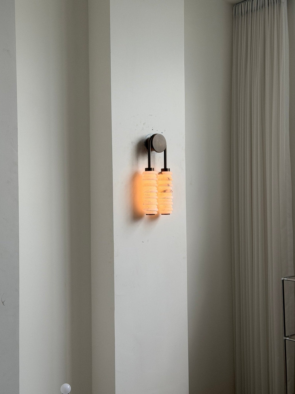 Distant view of a lit Clérachic sconce on a tall white wall beside a sheer curtain, giving a sense of scale.