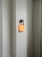Distant view of a lit Clérachic sconce on a tall white wall beside a sheer curtain, giving a sense of scale.