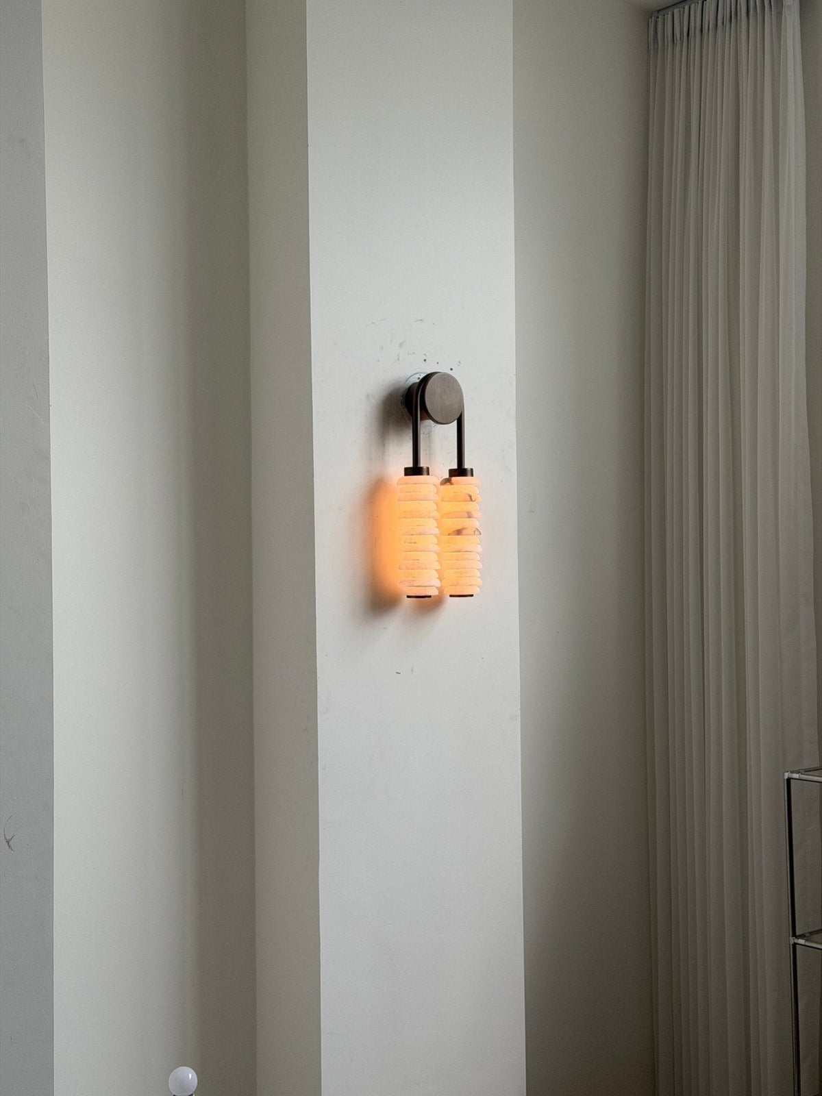 Distant view of a lit Clérachic sconce on a tall white wall beside a sheer curtain, giving a sense of scale.