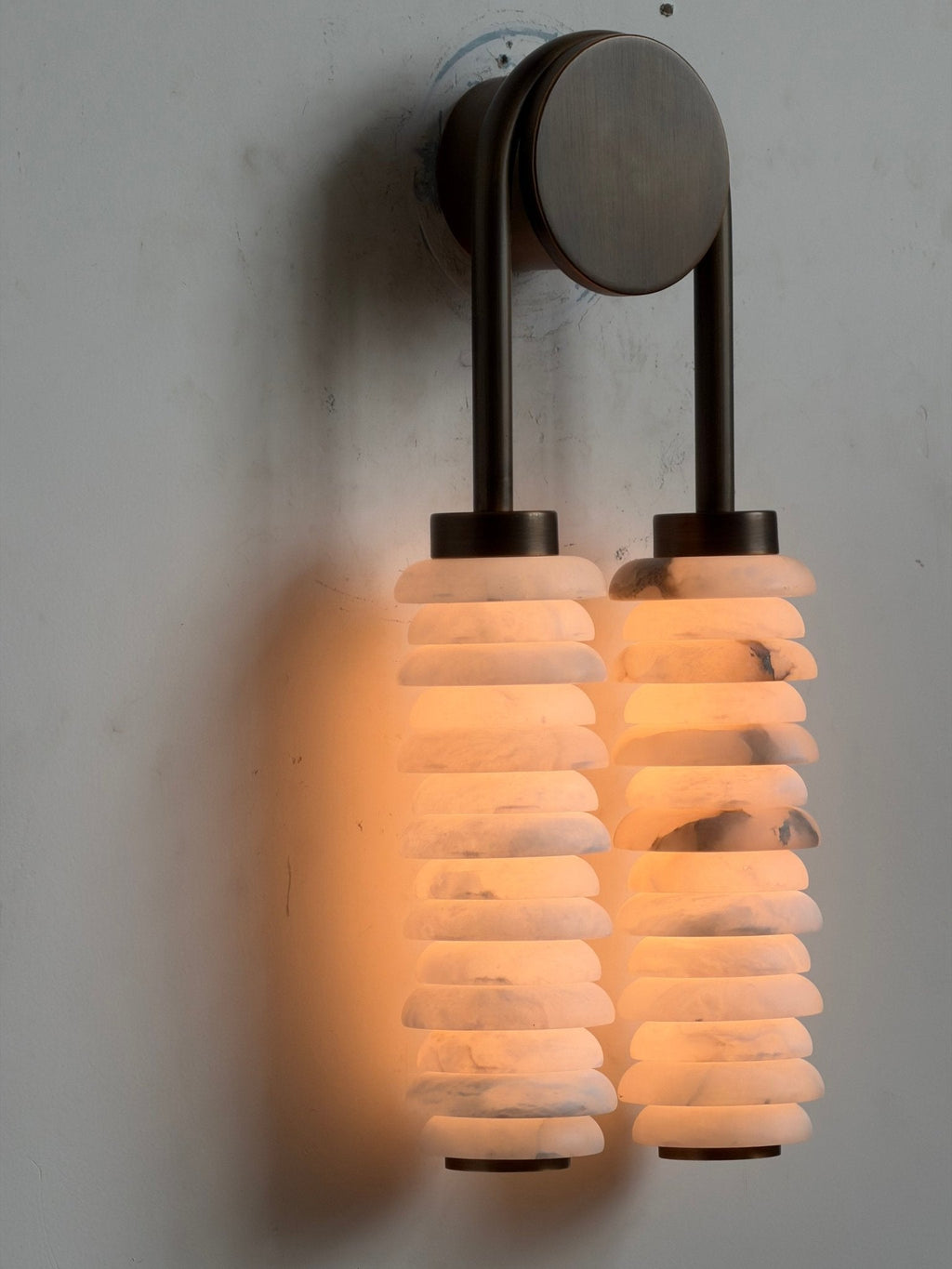 Clérachic sconce lit on a pale wall, the round bronze backplate and arch above two glowing alabaster columns.