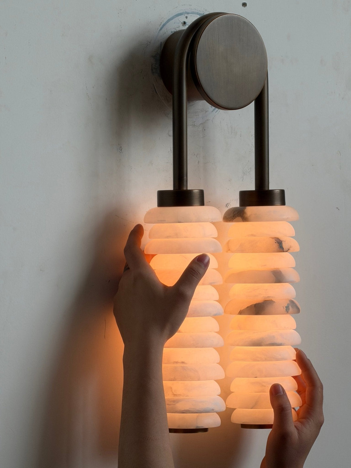 Hands near a lit Clérachic sconce, the veined alabaster discs glowing orange under the bronze arch.