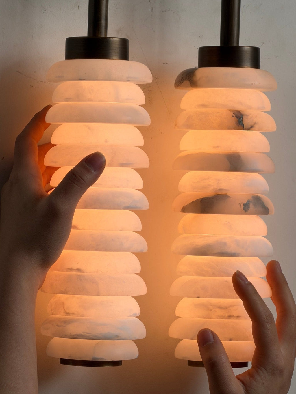 Close view of hands around the two glowing alabaster columns of a Clérachic sconce, with bronze caps above.