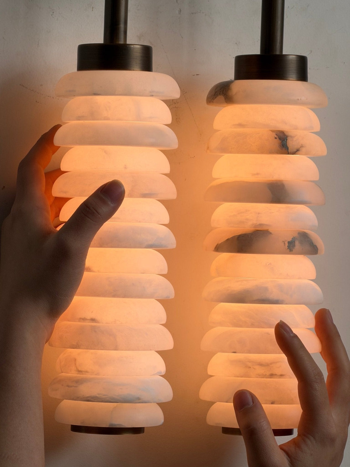 Close view of hands around the two glowing alabaster columns of a Clérachic sconce, with bronze caps above.