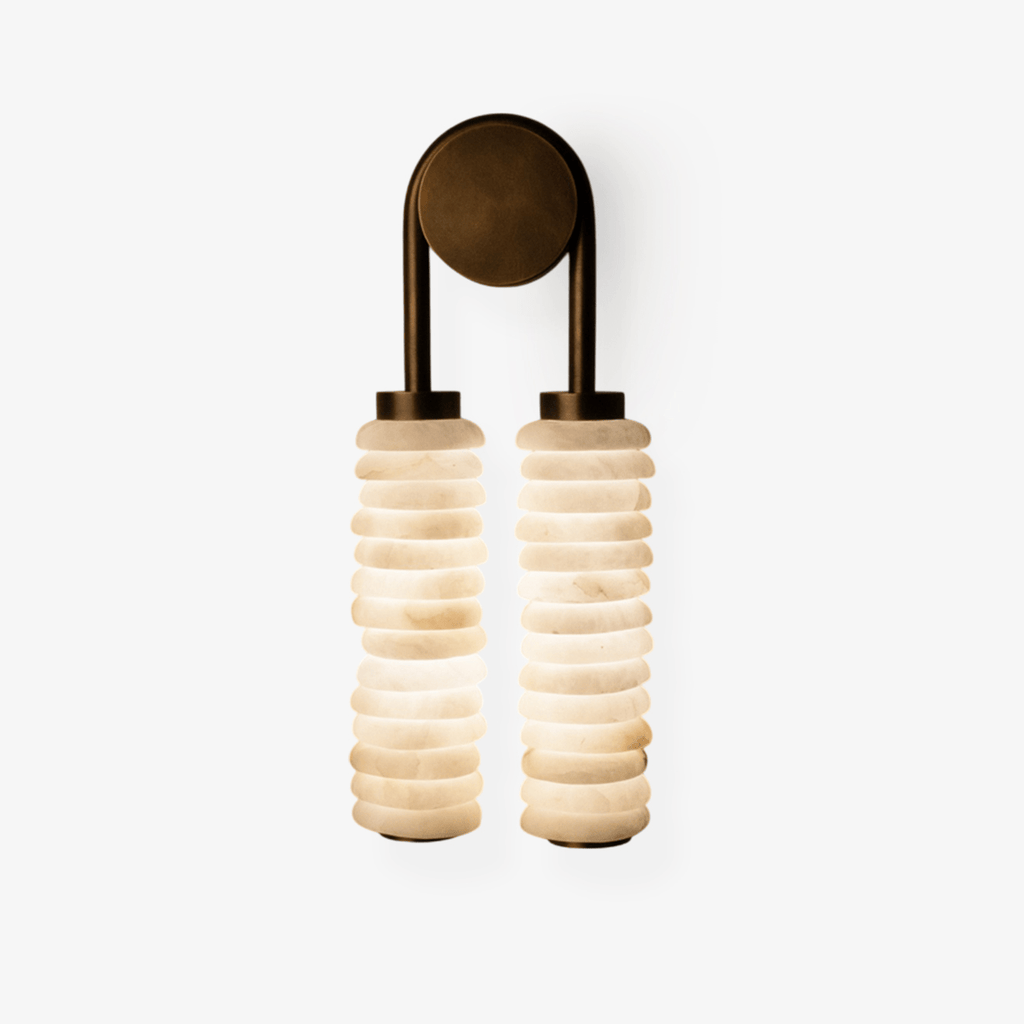 Clérachic wall sconce lit on a white backdrop, two alabaster columns glowing soft white under a bronze arch.
