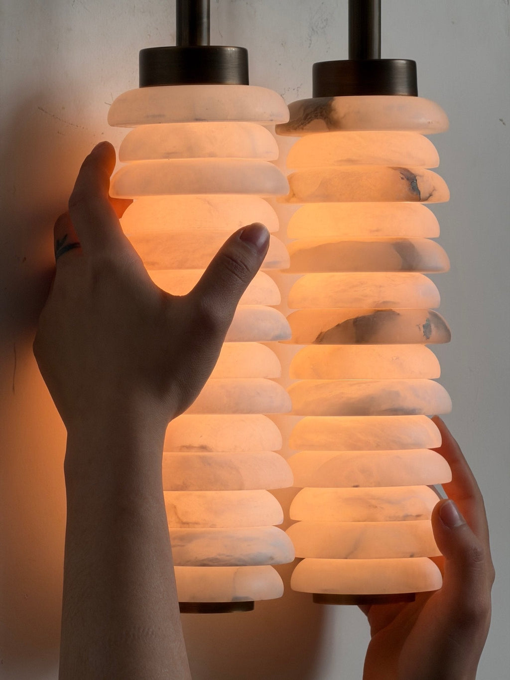 Hands on a lit Clérachic sconce, close in, showing the alabaster discs glowing orange with grey veining.