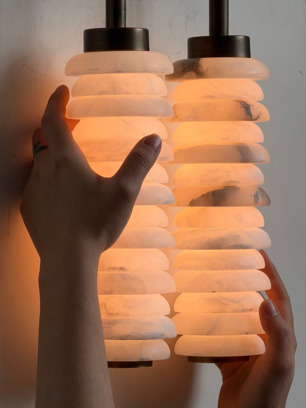 Hands on a lit Clérachic sconce, close in, showing the alabaster discs glowing orange with grey veining.