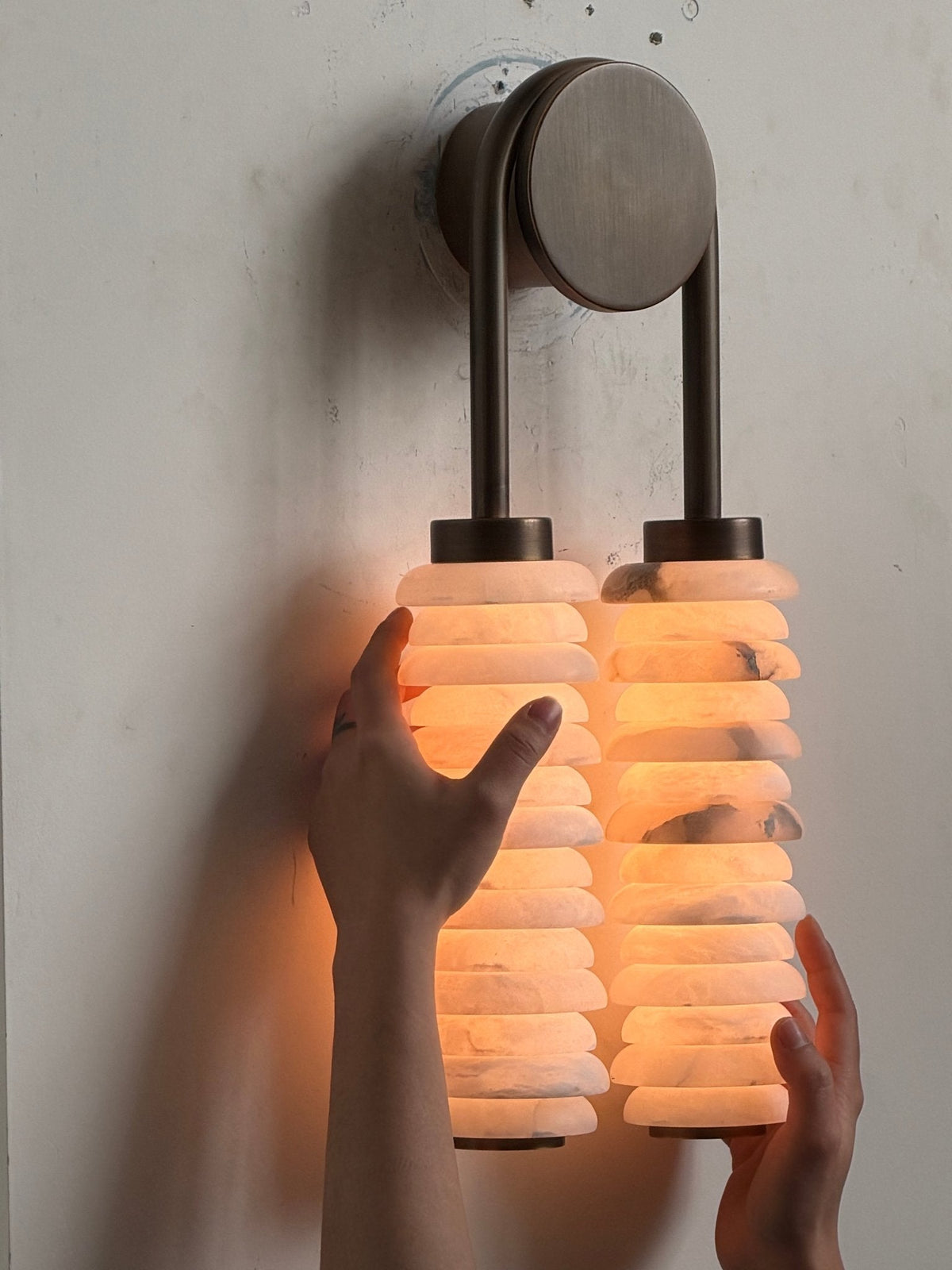 Lit Clérachic sconce on a plain wall with a hand held to one side, the bronze plate above two warm-glowing columns.