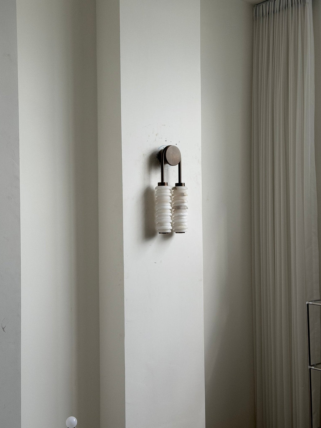 Distant view of an unlit Clérachic sconce on a white wall beside a curtain, small against the tall wall.