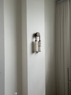 Distant view of an unlit Clérachic sconce on a white wall beside a curtain, small against the tall wall.