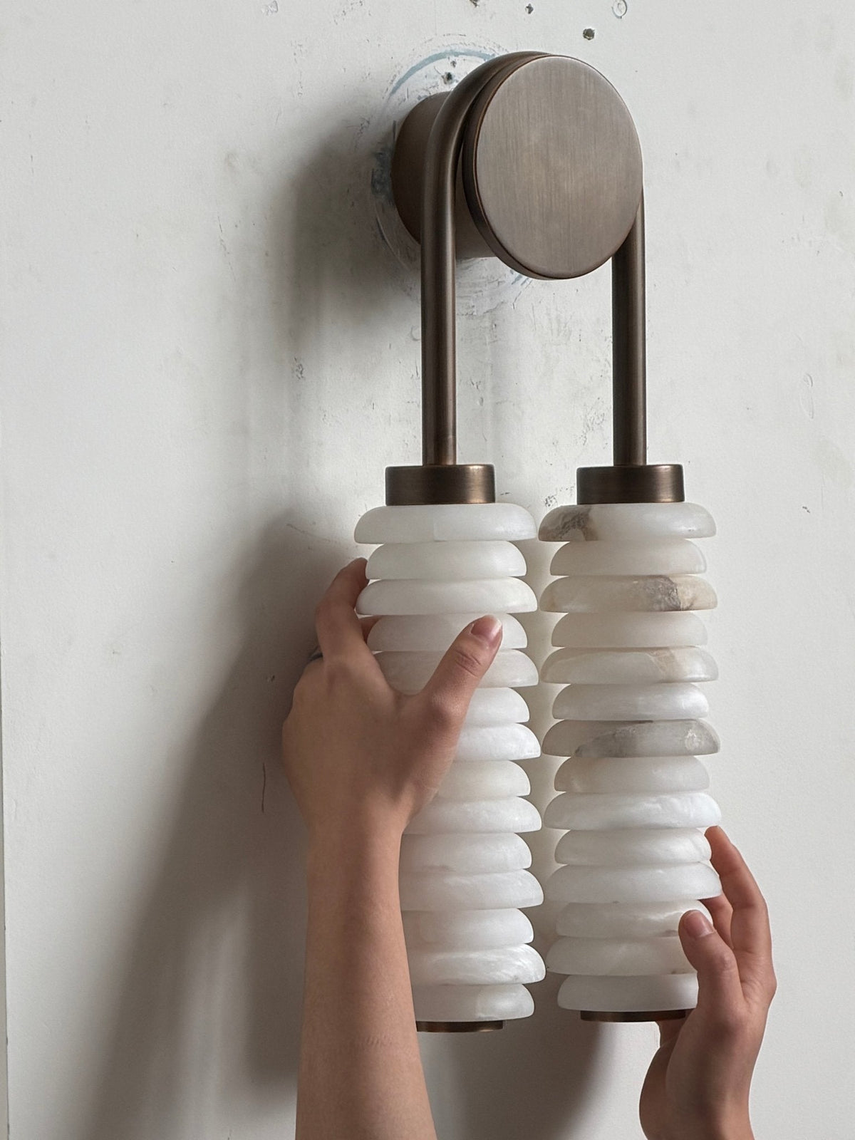 Clérachic sconce on a white wall with a hand on each column, the round bronze backplate above the alabaster discs.