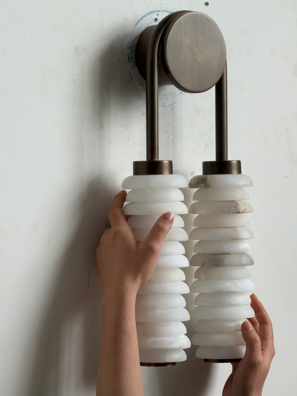 Hands resting against the unlit alabaster columns of a Clérachic sconce under a bronze arch and round backplate.