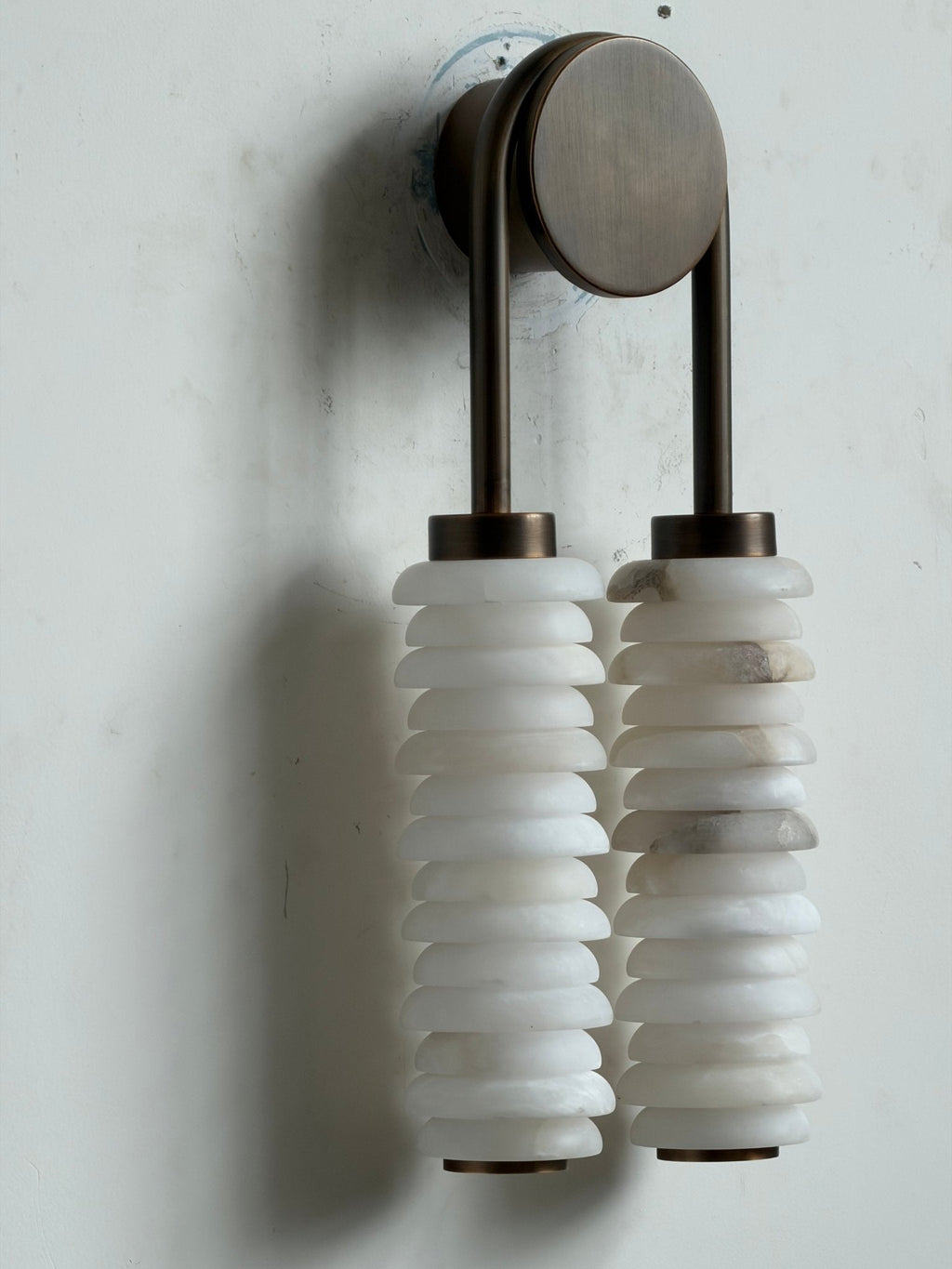 Unlit Clérachic sconce on a white wall, bronze arch and round plate above two veined alabaster columns.