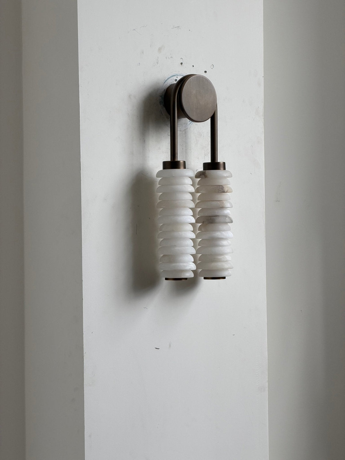 Clérachic sconce unlit on a white wall panel, the two alabaster columns hanging beneath a small bronze arch.