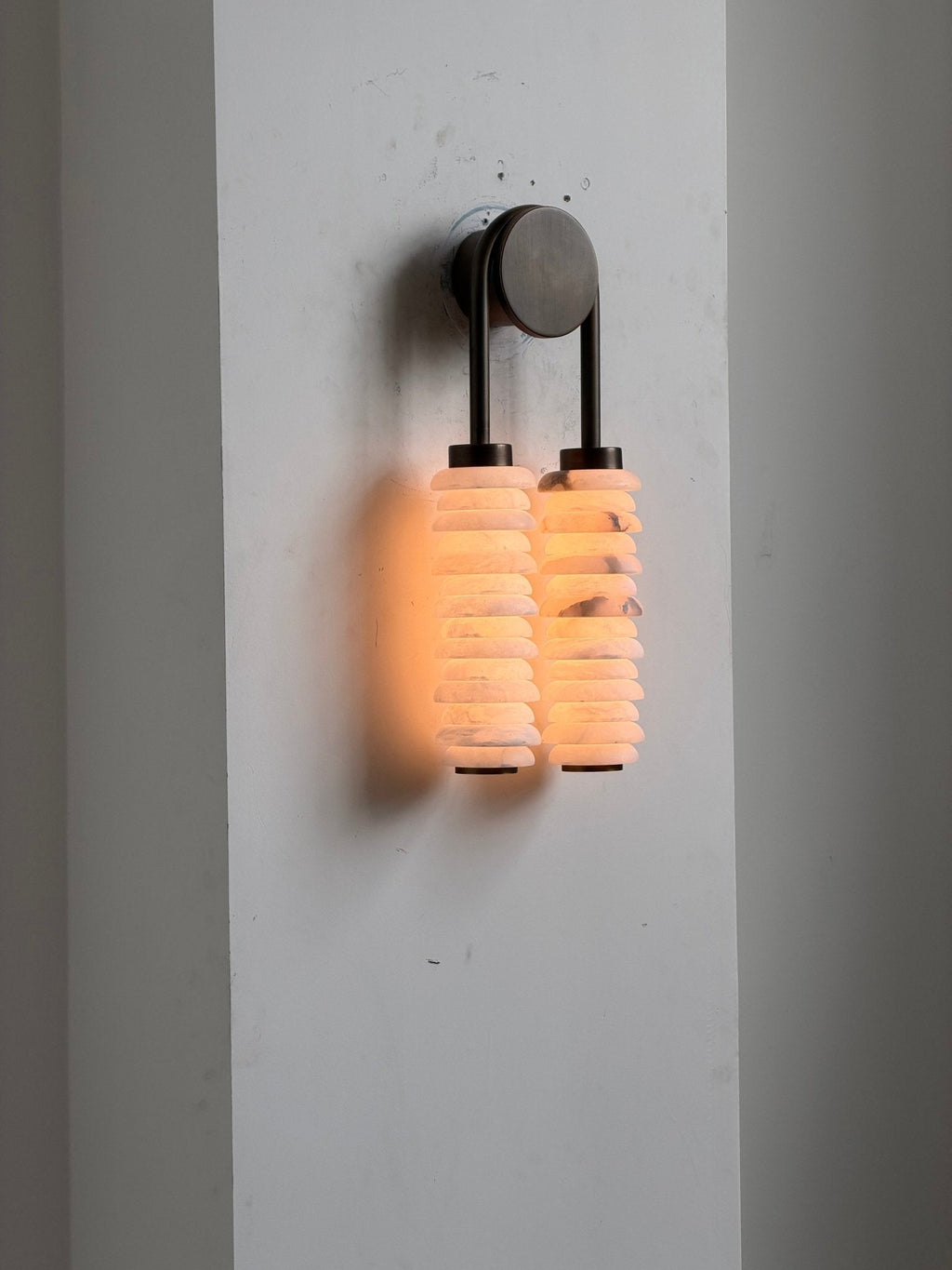Clérachic sconce on a white wall panel, switched on so the alabaster columns glow orange beneath the bronze arch.