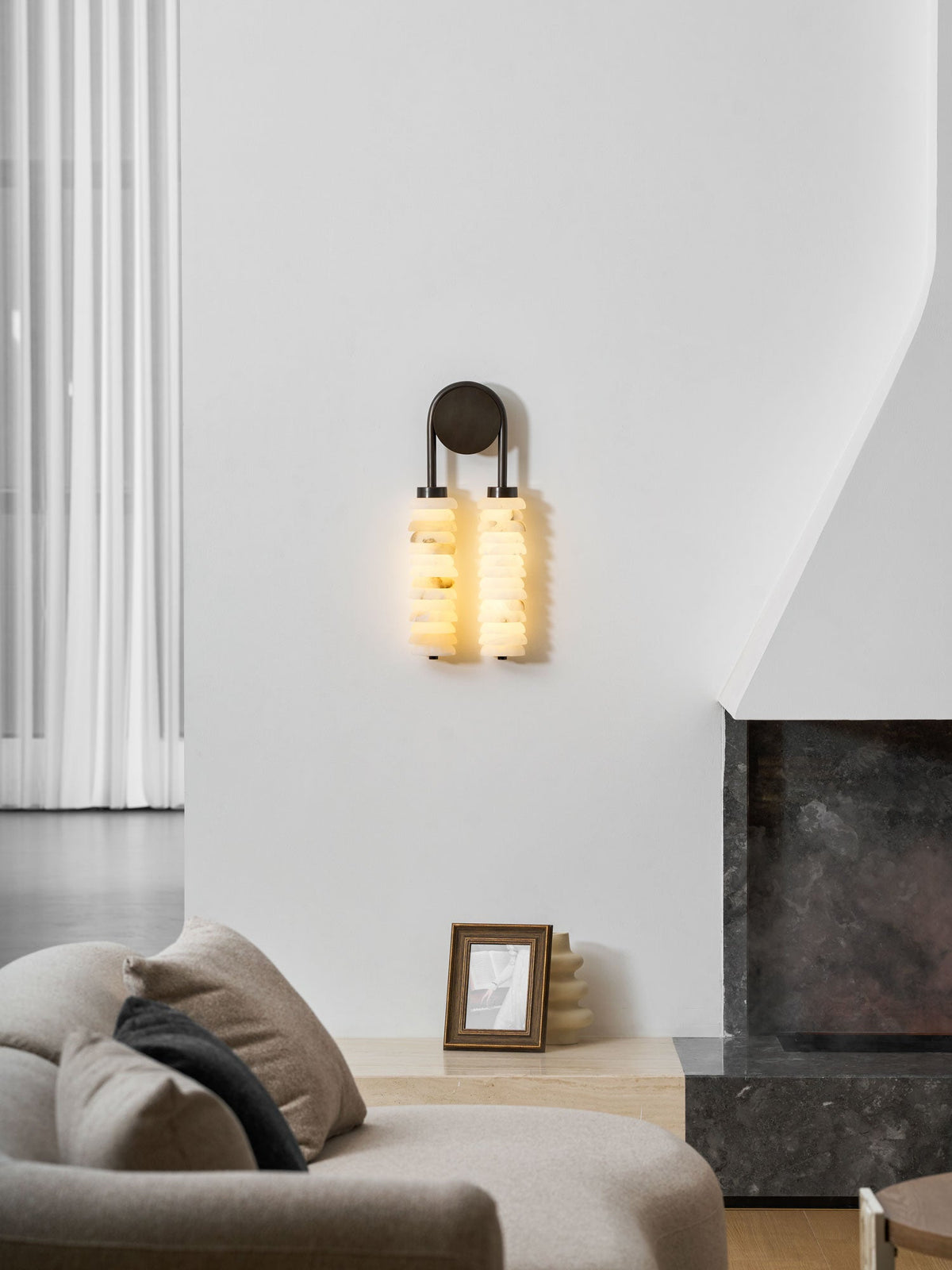 Lit Clérachic sconce on a white wall by a fireplace, with a cushioned sofa and a framed picture in the foreground.