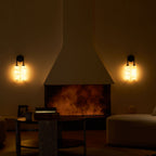 Two lit Clérachic sconces flank a glowing fireplace in a dim room, with a round table and a boucle pouffe.