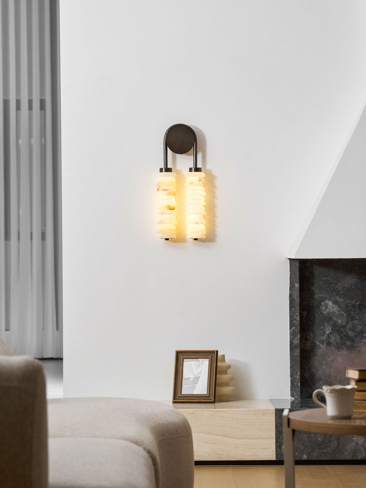Lit Clérachic sconce on a white wall, the alabaster glowing warm, above a framed picture, a sofa and a mug on a round table.