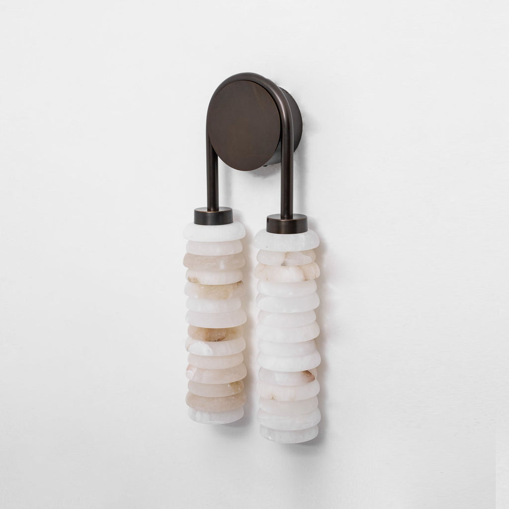 Clérachic sconce unlit on a pale wall, two columns of mixed cream and white alabaster under a dark bronze arch.