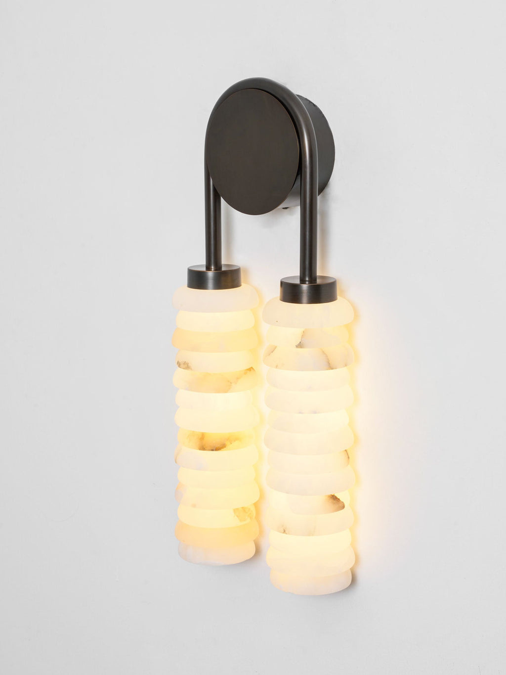 Lit Clérachic sconce in dark bronze on a pale wall, the cream and white alabaster discs glowing a warm yellow.