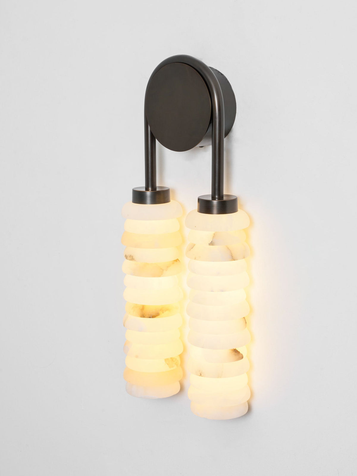 Lit Clérachic sconce in dark bronze on a pale wall, the cream and white alabaster discs glowing a warm yellow.