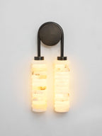 Lit Clérachic sconce with a dark bronze arch, the two alabaster columns glowing warm white against a pale grey wall.