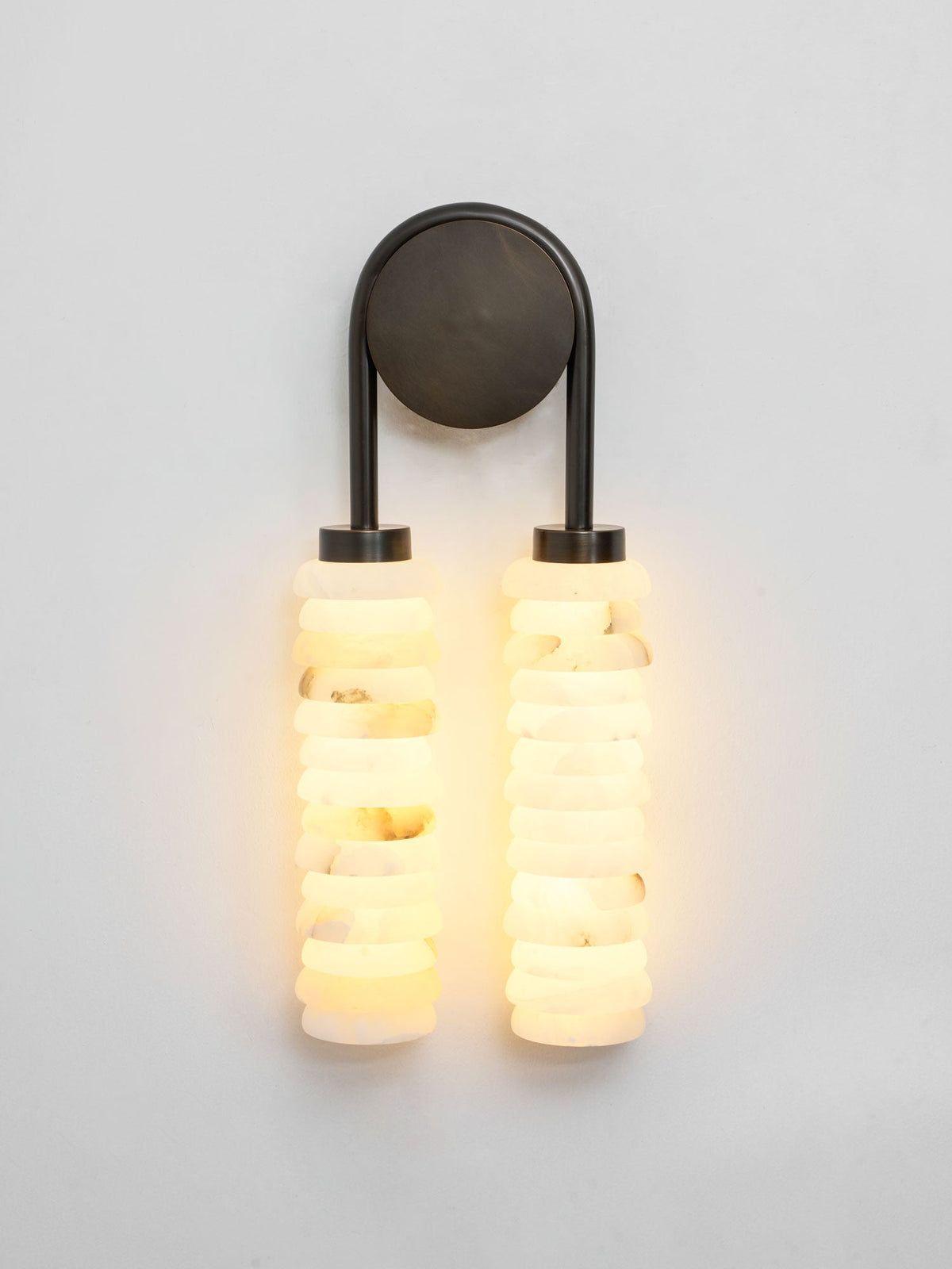 Lit Clérachic sconce with a dark bronze arch, the two alabaster columns glowing warm white against a pale grey wall.