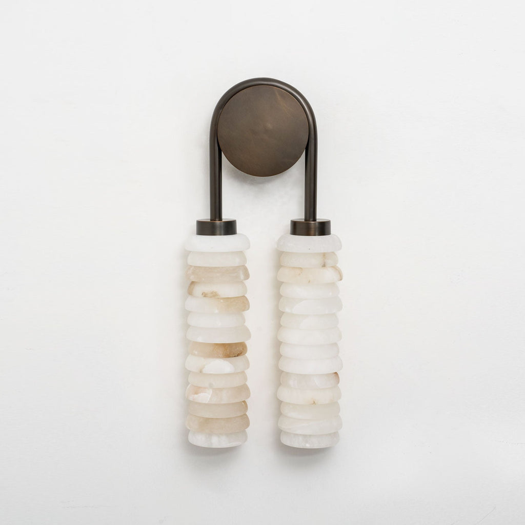 Clérachic sconce in dark bronze, unlit on a pale grey wall, with two stacked columns of cream and white alabaster.