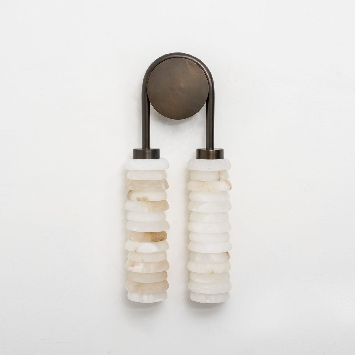 Clérachic sconce in dark bronze, unlit on a pale grey wall, with two stacked columns of cream and white alabaster.