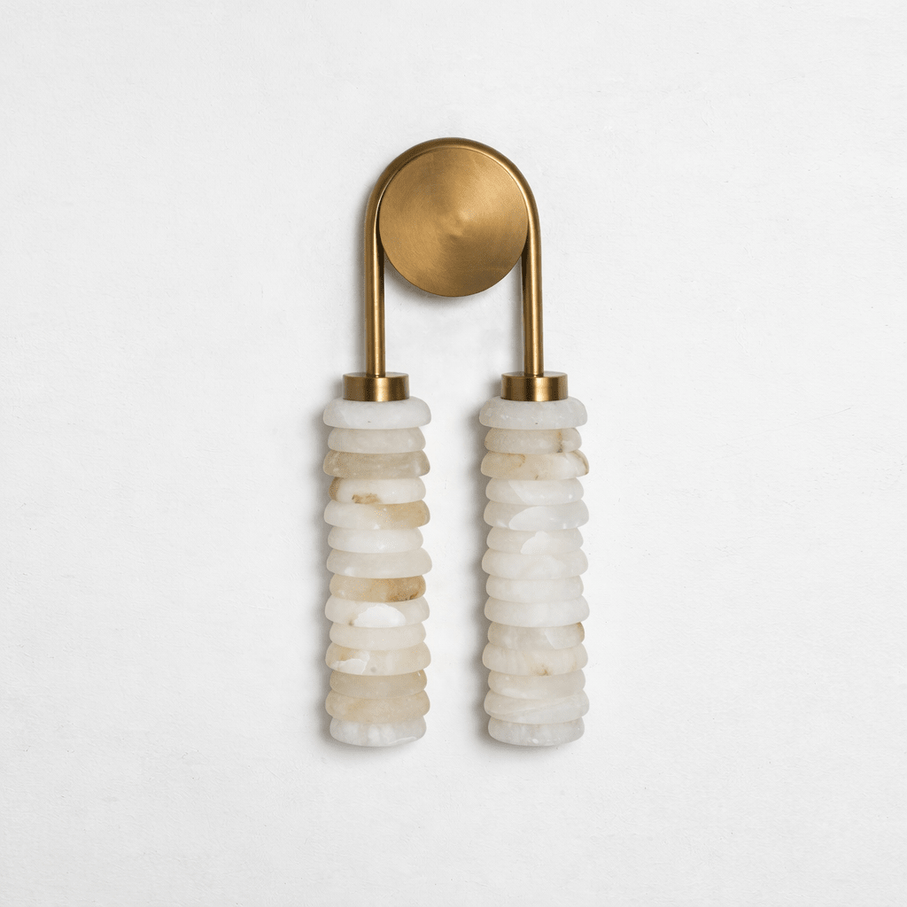Clérachic sconce with a brushed antique-brass arch and plate, unlit, over two columns of cream alabaster on a pale wall.
