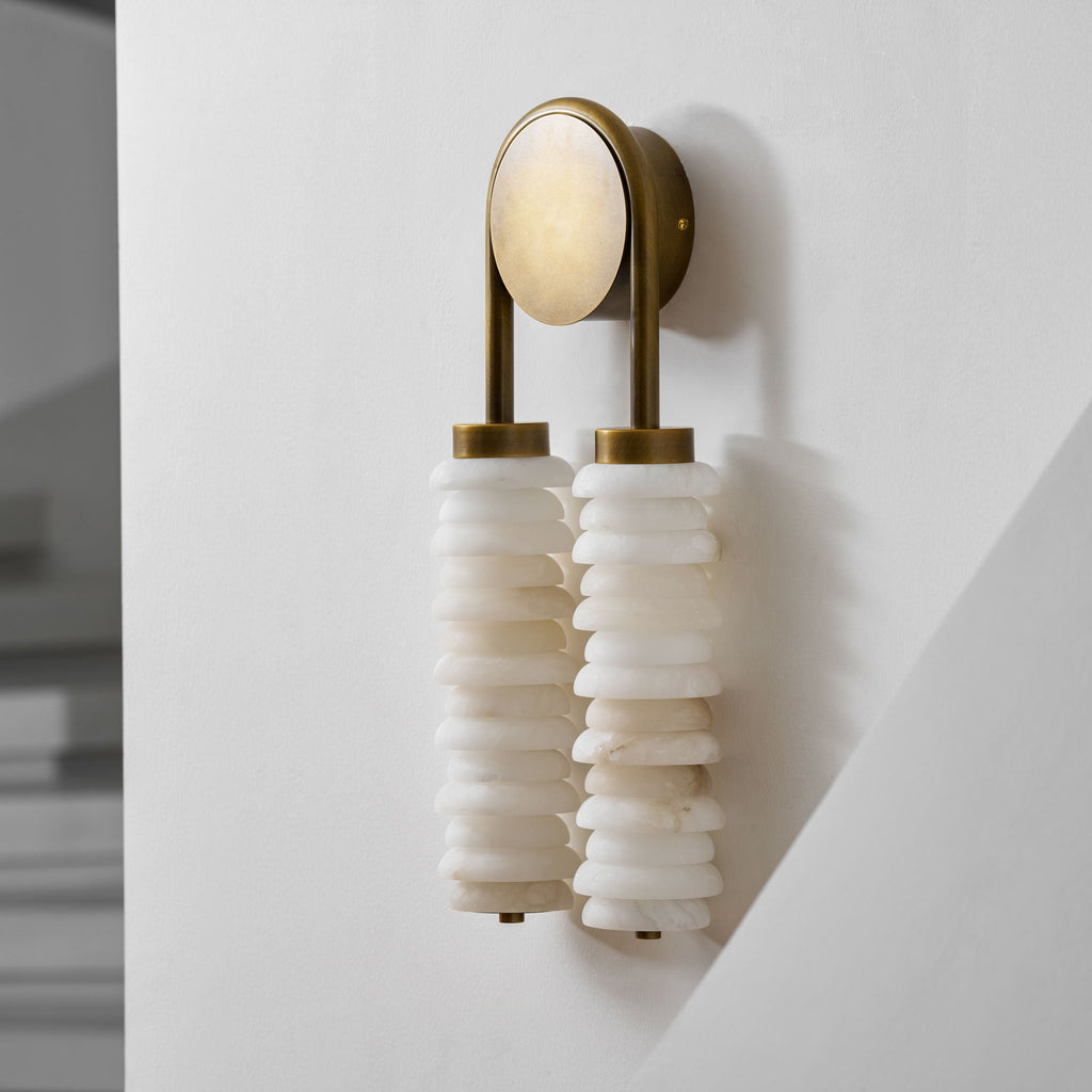 Angled view of a Clérachic sconce in antique brass on a white wall, with white alabaster discs and a pale round backplate.