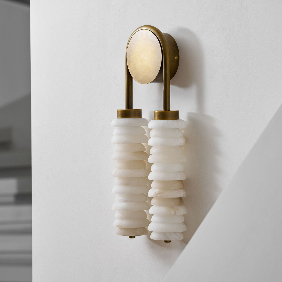 Angled view of a Clérachic sconce in antique brass on a white wall, with white alabaster discs and a pale round backplate.