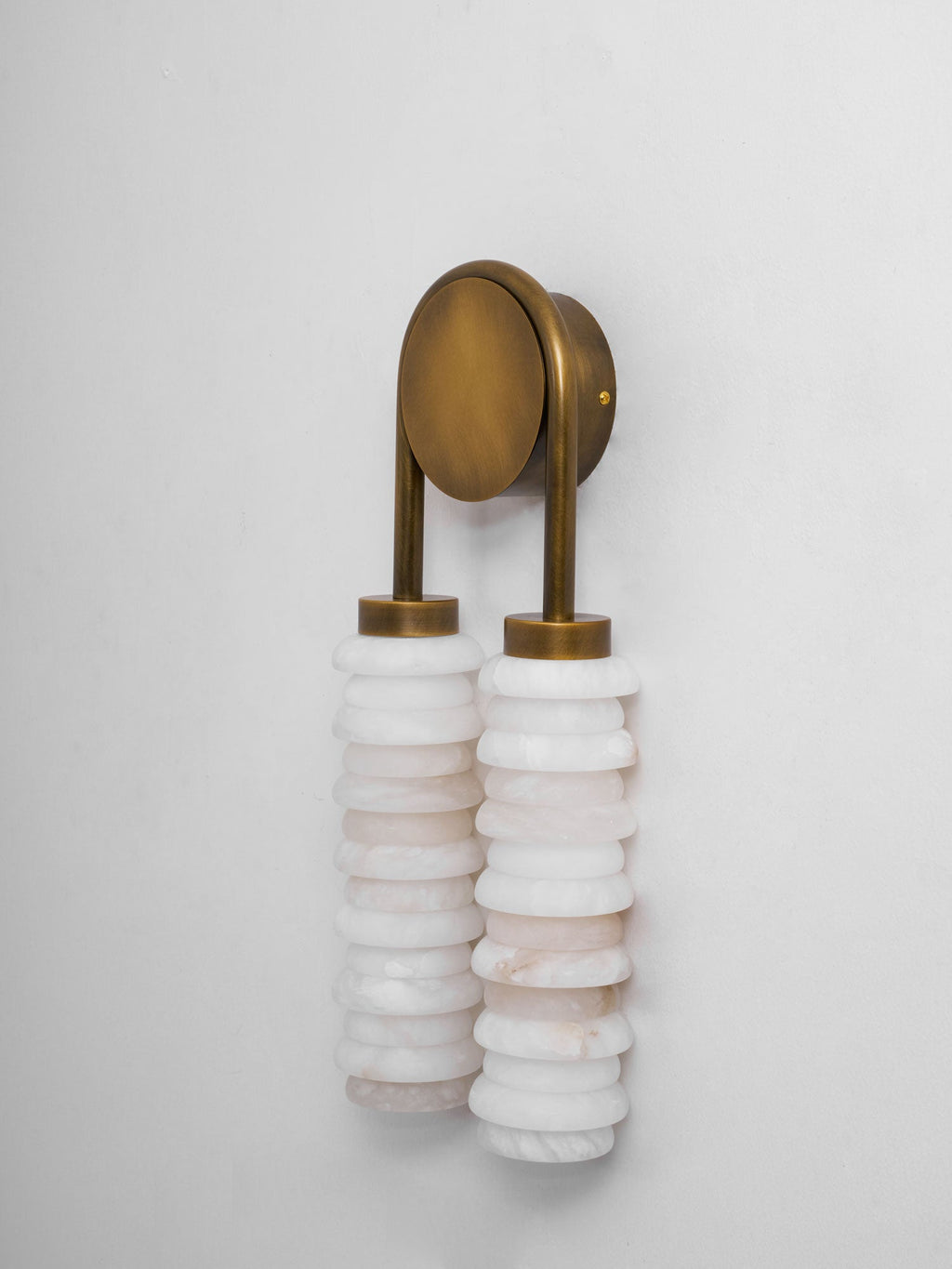 Clérachic sconce with an antique-brass arch and plate seen from the side, over two white alabaster columns on a pale wall.