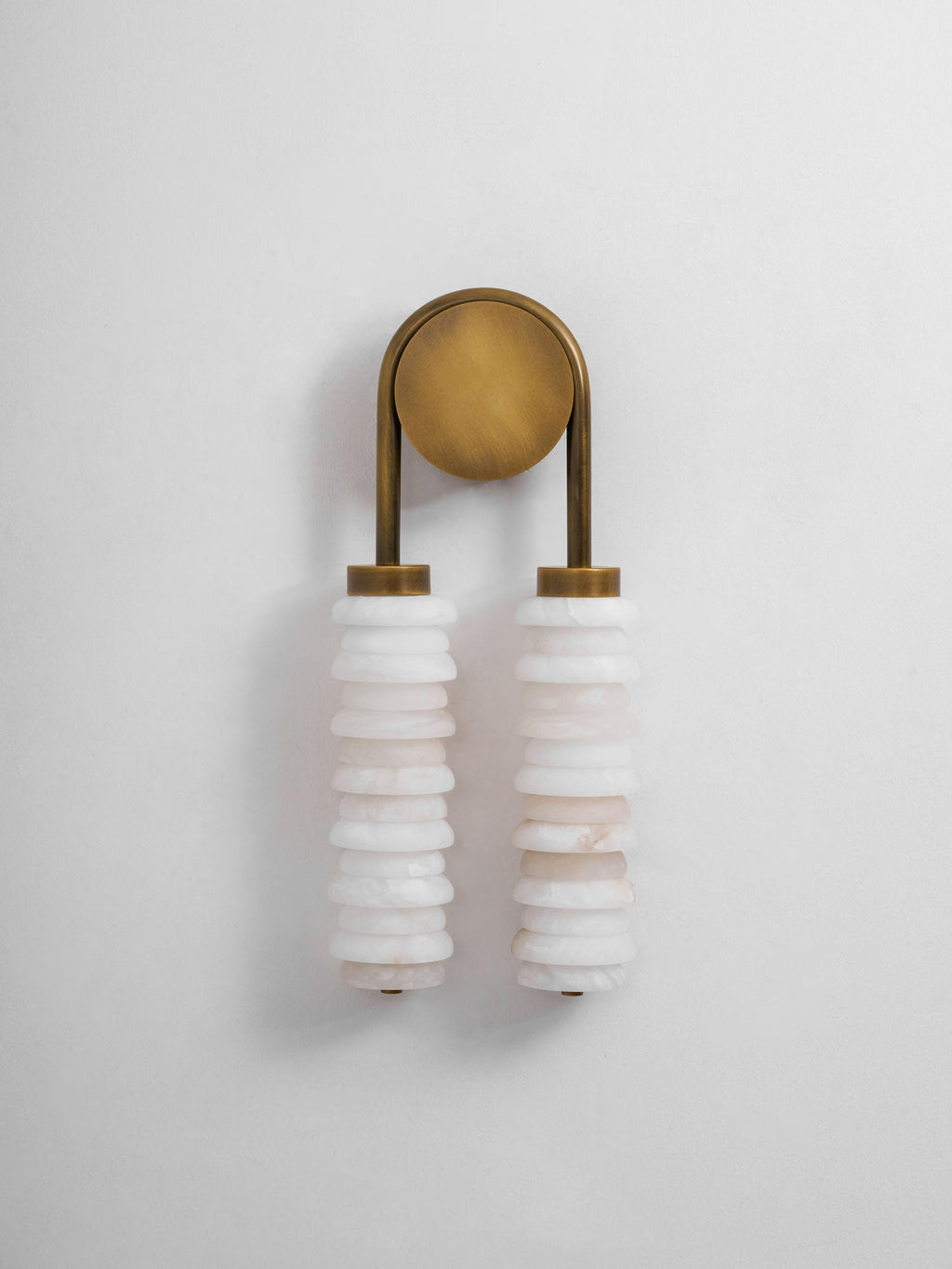 Small unlit Clérachic sconce in antique brass on a pale wall, two white alabaster columns hanging beneath the round plate.